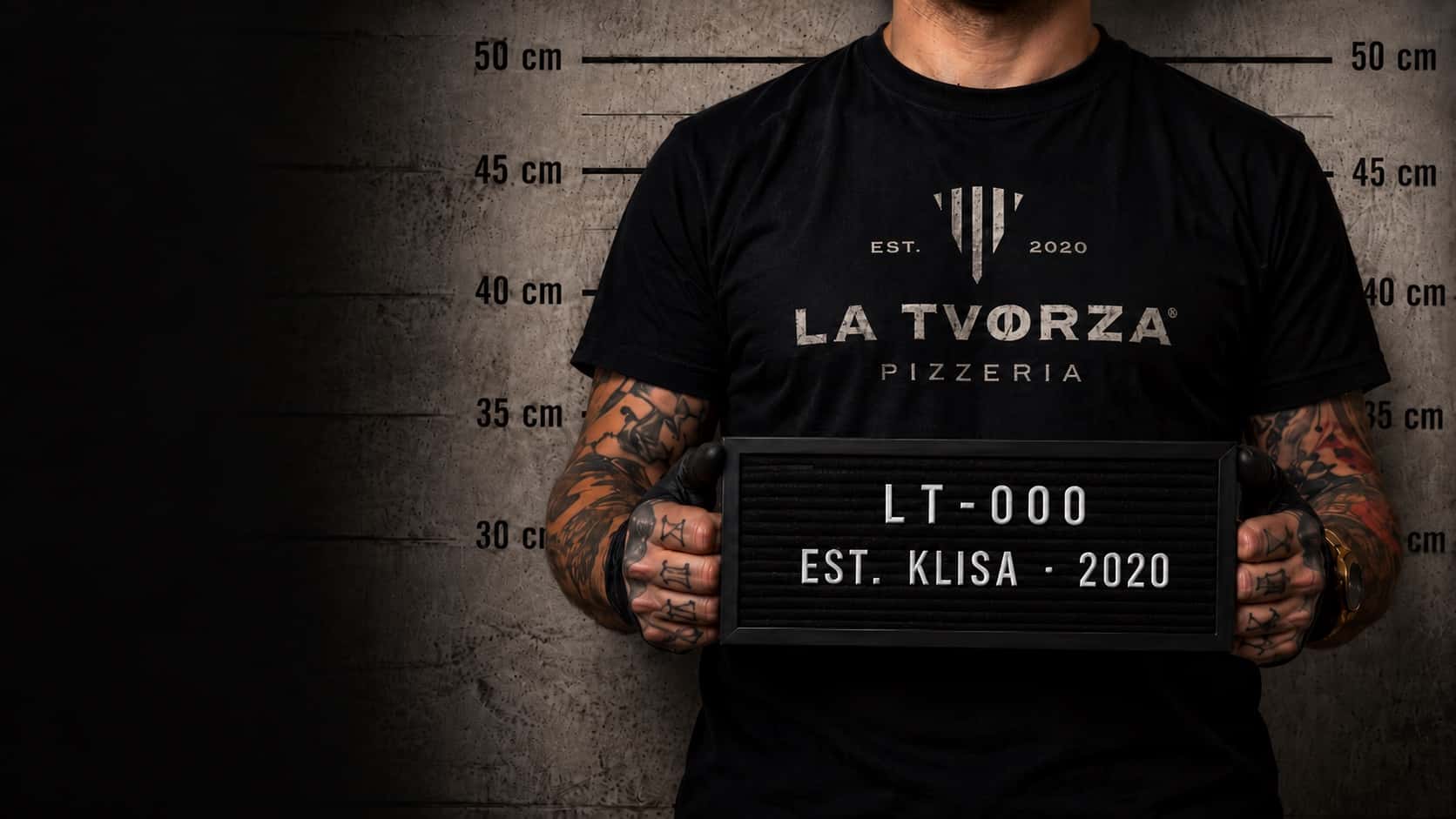 A pizzaiolo in a black La Tvorza t-shirt holds an LT-000, Est. Klisa 2020 placard in front of a height chart, like a police mugshot